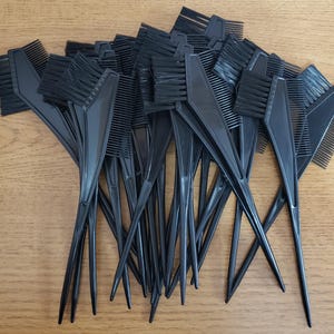 May include: A collection of black hair dye brushes and combs. Each brush has a comb on one side and a brush on the other. The handles are long and pointed. The items are arranged on a light brown wooden surface.