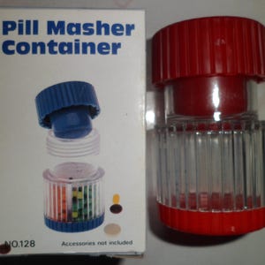 Könnte beinhalten: Ein roter und klarer Pillenzerkleinerer-Behälter mit blauem Deckel, neben einer Schachtel mit der Aufschrift "Pill Masher Container". Die Schachtel zeigt eine Explosionsansicht des Zerkleinerers. Der Zerkleinerer ist zum Zerkleinern von Pillen bestimmt.