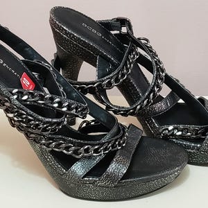 May include: Pair of BCBG Generation high-heeled sandals. The open-toe shoes are black with a textured finish and chain detailing. The heels are approximately 10 cm high.