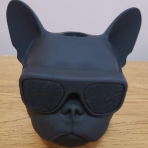 Può includere: Un altoparlante wireless nero opaco a forma di testa di bulldog francese. Il cane presenta grandi orecchie dritte e indossa occhiali da sole neri che coprono gli occhi. La griglia dell'altoparlante è integrata nelle lenti degli occhiali.