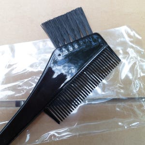 May include: A black hair dye applicator tool with a comb and brush. The comb has fine teeth, and the brush has black bristles. The tool is made of shiny black plastic and is designed for applying hair dye.