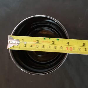 May include: A black, round container with a silver rim is measured with a yellow tape measure. The container is approximately 3 inches in diameter. The tape measure shows the measurement in inches.