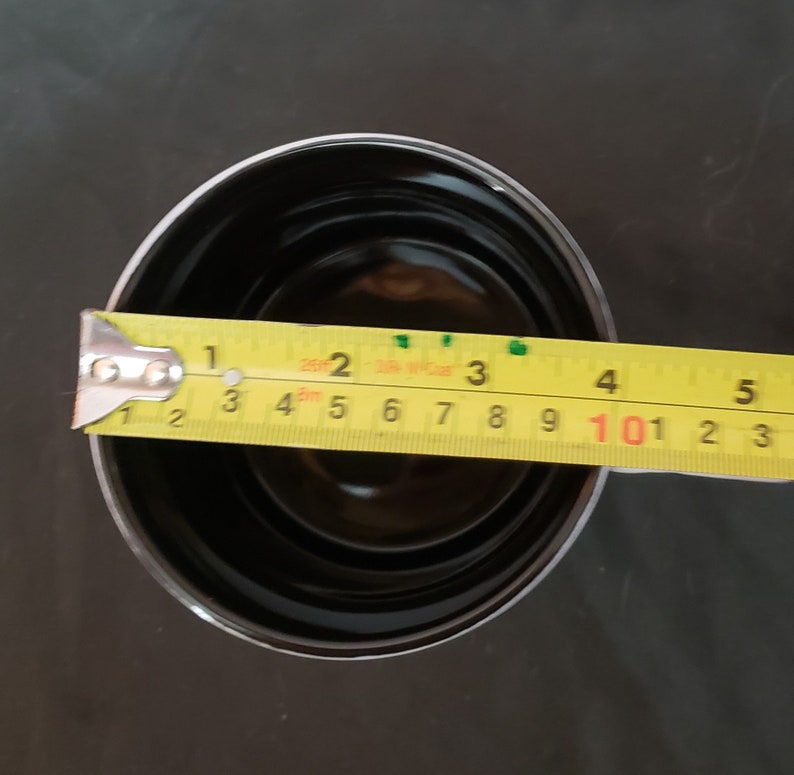 May include: A black, round container with a silver rim is measured with a yellow tape measure. The container is approximately 3 inches in diameter. The tape measure shows the measurement in inches.
