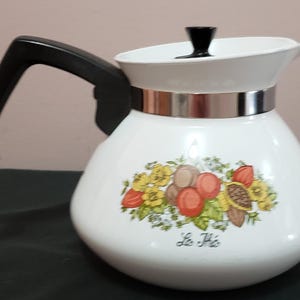 Peut inclure: Une théière blanche avec une anse et un couvercle noirs, dotée d'une bande métallique argentée autour du col. Le corps est orné d'une illustration botanique colorée de fruits et de fleurs, avec le texte manuscrit Le Thé sous le motif.