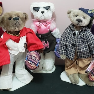 May include: A collection of six teddy bears dressed in various retro outfits. One bear wears a burgundy dress and hat, another a red and white jumpsuit, and a third sports sunglasses and a pink outfit. The bears are on display with tags.