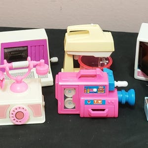 Pode incluir: Uma coleção de eletrodomésticos de brinquedo em miniatura em cores pastel. Inclui um toca-discos rosa com alto-falantes, um telefone branco e rosa, uma câmera de vídeo rosa, uma máquina de costura azul, um rádio rosa, um micro-ondas e um misturador.