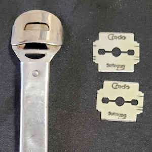Puede incluir: Una herramienta plateada para el cuidado de los pies con un soporte de cuchilla curvado y un mango largo. Se incluyen dos cuchillas de repuesto, cada una con el texto "Credo Solingen". Las cuchillas son blanquecinas y rectangulares.