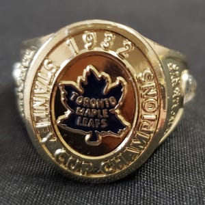 以下が含まれることがあります： 「1982」と「Stanley Cup Champions」の文字が入ったゴールドトーンのリング。リングには「Toronto Maple Leafs」の文字が入った青いカエデの葉のエンブレムが付いています。スポーツの収集品です。
