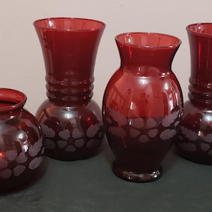 Könnte beinhalten: Vier rubinrote Glasvasen in verschiedenen Formen. Zwei Vasen haben einen bauchigen Boden und eine ausgestellte Oberseite, während die anderen beiden eine rundere Form haben. Jede Vase hat ein geätztes Blumendesign.