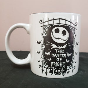 May include: White ceramic mug featuring a black graphic of Jack Skellington from The Nightmare Before Christmas. The design includes a gate, bats, and the text "THE MASTER OF FRIGHT". The mug has a curved handle and a black interior.