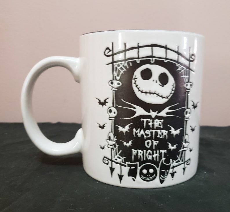 May include: White ceramic mug featuring a black graphic of Jack Skellington from The Nightmare Before Christmas. The design includes a gate, bats, and the text "THE MASTER OF FRIGHT". The mug has a curved handle and a black interior.