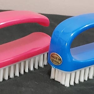 Op de afbeelding: Twee nagelborstels, één roze en één blauwe, met witte borstelharen en gebogen handvatten. Elke borstel heeft een rechthoekige basis en een handvat voor een gemakkelijke grip. De borstels zijn gemarkeerd met "Made in China".
