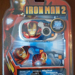 Puede incluir: Una cámara digital de Iron Man 2 en un paquete de plástico transparente. La cámara tiene un diseño azul y blanco con la cara de Iron Man. El paquete incluye el texto "Comes with 3 Changing Faceplates" y "Image Lab Software".