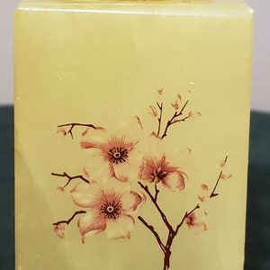 May include: A square, pale yellow alabaster box with a silver lid. The box has a floral design painted on the front, featuring pink flowers with brown stems and leaves.