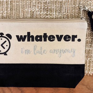 May include: A beige canvas makeup bag with a black zipper and a black strap. The bag has a black alarm clock graphic and the text "whatever. i'm late anyway" in silver glitter.