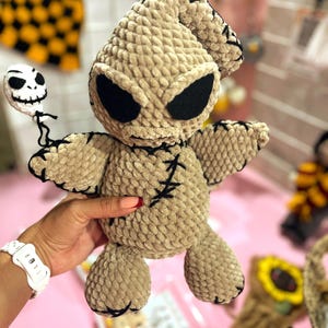 Puede incluir: Un juguete de peluche tejido a crochet del personaje Oogie Boogie de la película Pesadilla antes de Navidad. El juguete es beige con ojos negros y una boca cosida. Sostiene una pequeña calavera de crochet con una cara blanca.
