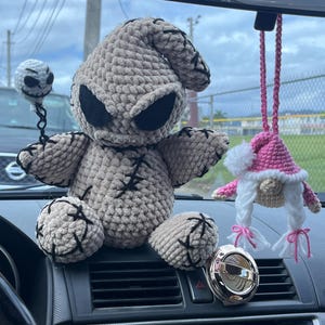 K&ouml;nnte beinhalten: Ein graues, geh&auml;keltes Pl&uuml;schtier, das einem Charakter aus dem Film "Nightmare Before Christmas" &auml;hnelt, sitzt auf dem Armaturenbrett eines Autos. Ein rosa geh&auml;kelter Zwerg, der am R&uuml;ckspiegel h&auml;ngt, ist im Hintergrund zu sehen.