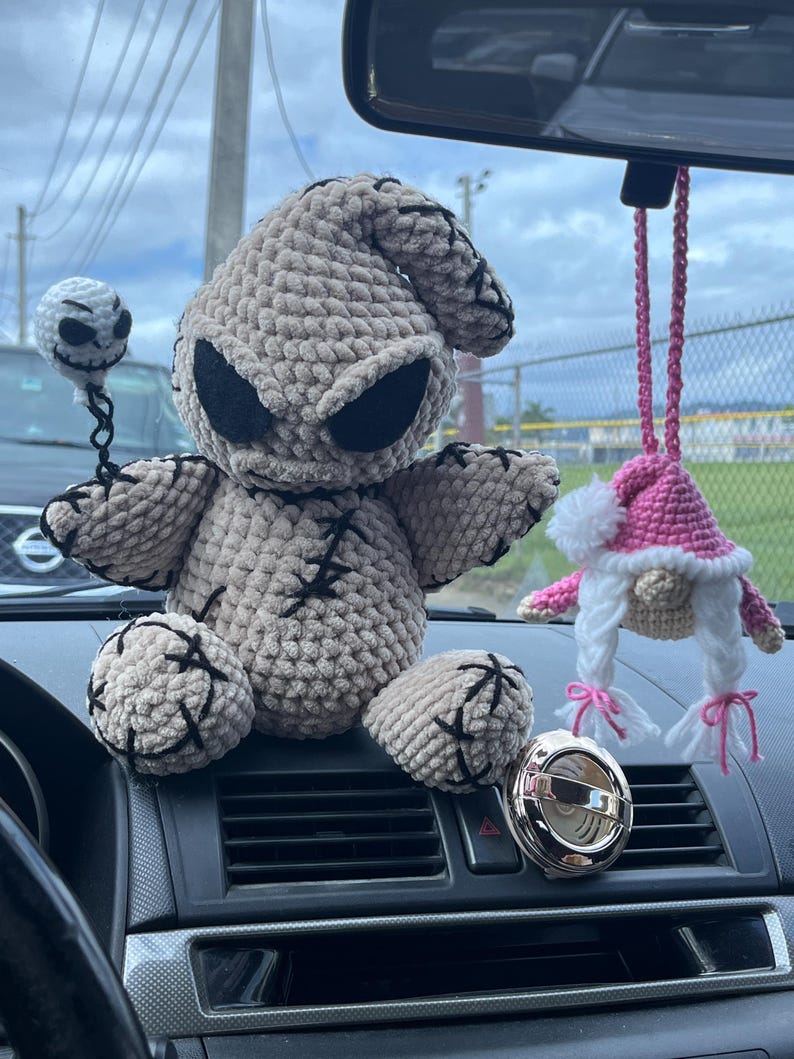 K&ouml;nnte beinhalten: Ein graues, geh&auml;keltes Pl&uuml;schtier, das einem Charakter aus dem Film "Nightmare Before Christmas" &auml;hnelt, sitzt auf dem Armaturenbrett eines Autos. Ein rosa geh&auml;kelter Zwerg, der am R&uuml;ckspiegel h&auml;ngt, ist im Hintergrund zu sehen.