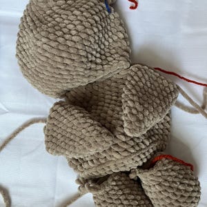K&ouml;nnte beinhalten: Ein braunes, geh&auml;keltes Stofftier in Form eines Hundes. Der Hund hat eine lange Schnauze, Schlappohren und einen Schwanz. Der Hund liegt auf der Seite mit ausgestreckten Beinen. Der Hund ist aus einem weichen, flauschigen Material gefertigt. Der Hund hat rotes Garn daran befestigt.