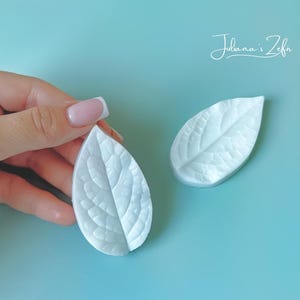 Puede incluir: Dos decoraciones blancas en forma de hoja con vetas detalladas, una de ellas sostenida por una mano con manicura francesa. Están sobre un fondo azul claro. La imagen incluye el texto "Juliana's Zefa".