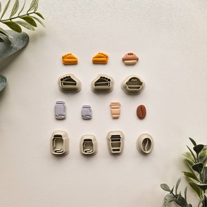 May include: A set of 12 cookie cutters in various shapes, including pies, coffee cups, jars, and coffee beans. The cutters are made of white plastic and are arranged in rows on a white surface.