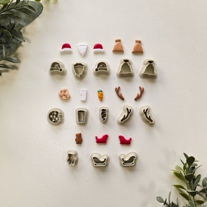 May include: A set of 20 Christmas-themed cookie cutters, including Santa hats, reindeer antlers, a gingerbread man, a stocking, and a milk glass. The cutters are made of white plastic and are arranged on a white background.