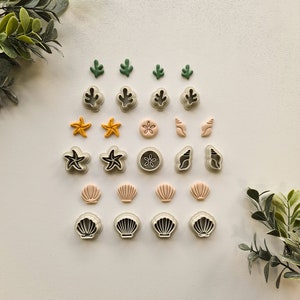 May include: A selection of sea-themed cookie cutters, featuring starfish, seashells, and sand dollars. The cutters are in shades of green, yellow, and pink, with white and black outlines. The arrangement is on a white surface, with green foliage in the corners.