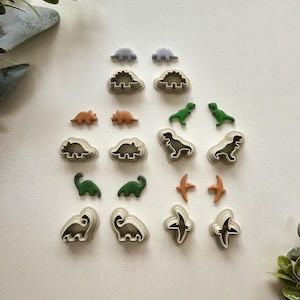 May include: A set of 12 dinosaur cookie cutters in various shapes and sizes. The cutters are made of plastic and are in shades of green, orange, and grey.