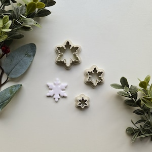 May include: Four snowflake cookie cutters of varying sizes on a white background. The largest cutter is in the top left corner, and the smallest cutter is in the bottom right corner. The cutters are made of a light grey plastic.