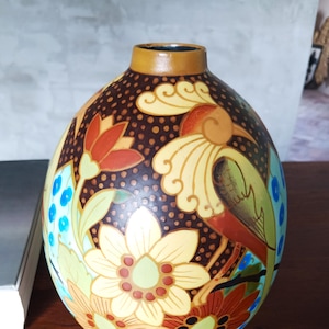 Op de afbeelding: Een ronde keramische vaas met een donkerbruine achtergrond met een gestippelde textuur. Het oppervlak is versierd met kleurrijke illustraties van grote bloemen in tinten oranje, geel en rood, naast een gestileerde vogel en blauwe bessen.