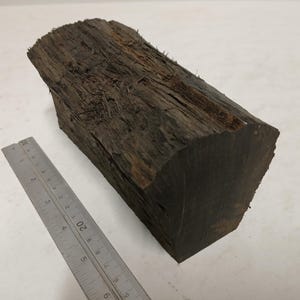 Puede incluir: Un trozo de madera en bruto de color marrón oscuro con una textura rugosa y una superficie astillada. Una regla metálica está colocada junto a la madera, mostrando que la longitud de la pieza es de unos 14 centímetros en la escala métrica.