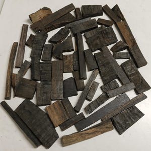 May include: A collection of numerous small, irregular pieces of dark brown wood with visible grain and rough, weathered textures. The fragments vary in length and width, featuring straight and angled edges, scattered across the frame.