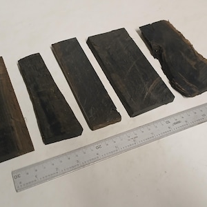 May include: Five dark wood planks of varying sizes, arranged on a white surface. A silver ruler with metric measurements is placed next to the planks. The wood has a dark brown color with visible grain.