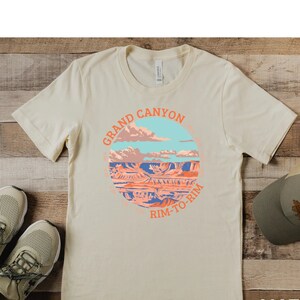 May include: A beige t-shirt with a circular graphic of the Grand Canyon with the text "Grand Canyon Rim-To-Rim" in orange. The t-shirt is on a wooden surface with a pair of beige sneakers and a brown baseball cap.