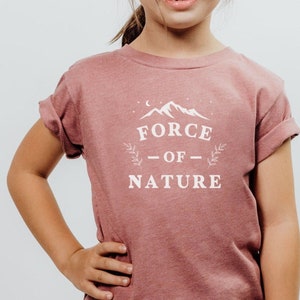 May include: A young person wearing a pink t-shirt with the text "FORCE OF NATURE" in white. The shirt features a white mountain graphic and stars. The person is wearing denim shorts with lace trim.