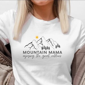 May include: White t-shirt with a black and white graphic of mountains, a sun, and trees. The text "Mountain Mama enjoying the great outdoors" is printed on the shirt.