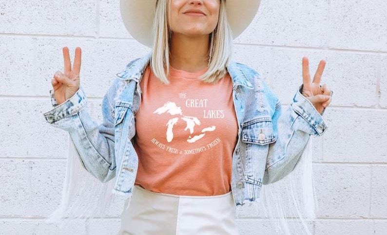 May include: A woman wearing a peach colored t-shirt with a white graphic of the Great Lakes and the text "Always Fresh - Sometimes Frozen". She is also wearing a light blue denim jacket with white fringe and a white hat.