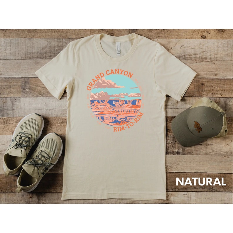 May include: A beige t-shirt with a circular graphic of the Grand Canyon with the text "Grand Canyon Rim-To-Rim" in orange. The t-shirt is on a wooden surface with a pair of beige sneakers and a brown baseball cap.