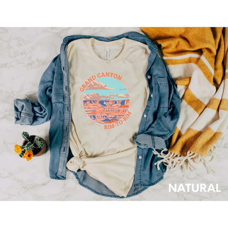 May include: A beige t-shirt with a graphic of the Grand Canyon and the text "Grand Canyon Rim-to-Rim" in a circle. The t-shirt is laid out on a marble surface with a blue denim shirt and a striped blanket.
