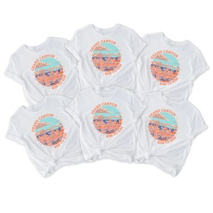 May include: Six white t-shirts with a graphic print of the Grand Canyon. The graphic features a colorful illustration of the canyon with the text "Grand Canyon" and "Rim-to-Rim" in a circle.
