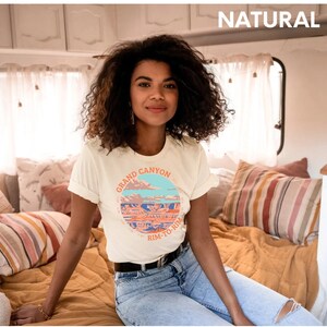 May include: A white t-shirt with a graphic of the Grand Canyon and the text "Grand Canyon Rim-to-Rim". The model is wearing the shirt and sitting on a bed.