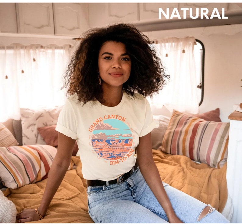 May include: A white t-shirt with a graphic of the Grand Canyon and the text "Grand Canyon Rim-to-Rim". The model is wearing the shirt and sitting on a bed.