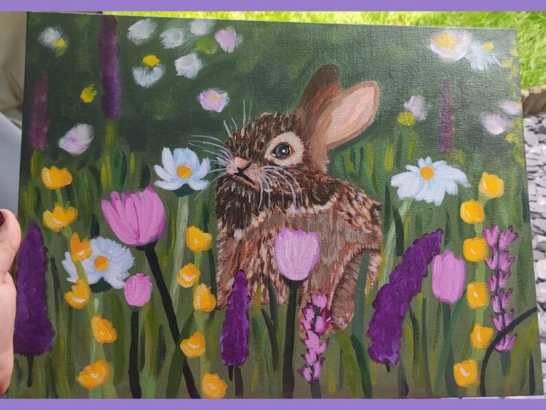 May include: An acrylic painting on canvas featuring a brown rabbit centered in a lush green meadow filled with colorful wildflowers. The scene includes purple lavender stalks, bright yellow blossoms, white daisies, and soft pink tulip-like flowers.
