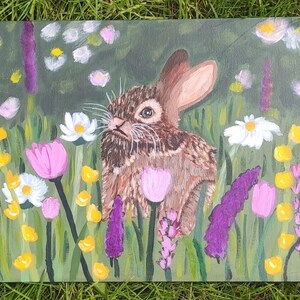 May include: An acrylic painting on canvas depicting a brown rabbit sitting in a field of colorful flowers. The scene features purple lupines, pink tulips, white daisies, and small yellow blossoms set against a green background of grass.
