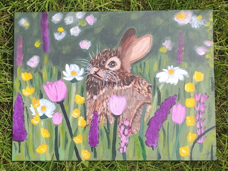 May include: An acrylic painting on canvas depicting a brown rabbit sitting in a field of colorful flowers. The scene features purple lupines, pink tulips, white daisies, and small yellow blossoms set against a green background of grass.