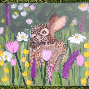 May include: A rectangular canvas painting featuring a brown rabbit centered among a field of colorful flowers. The scene includes white daisies, pink tulips, and tall purple stalks of lavender set against a green background with yellow accents.