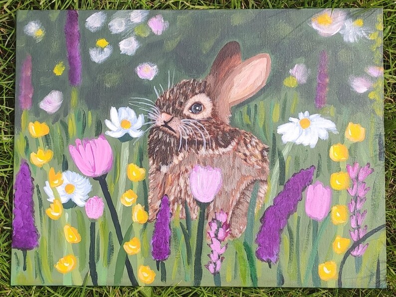 May include: A rectangular canvas painting featuring a brown rabbit centered among a field of colorful flowers. The scene includes white daisies, pink tulips, and tall purple stalks of lavender set against a green background with yellow accents.