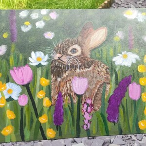 May include: An acrylic painting on canvas depicting a brown rabbit centered in a lush green meadow. The scene is filled with colorful wildflowers, including white daisies, pink tulips, and tall purple spikes, all rendered with visible brushstrokes.