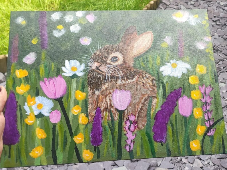 May include: An acrylic painting on canvas depicting a brown rabbit centered in a lush green meadow. The scene is filled with colorful wildflowers, including white daisies, pink tulips, and tall purple spikes, all rendered with visible brushstrokes.