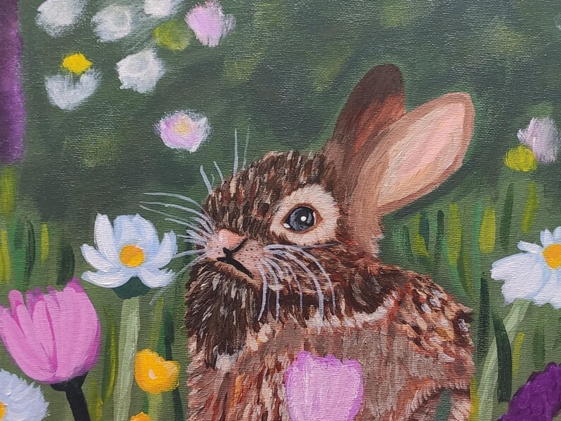 May include: An oil painting of a brown rabbit with long ears and white whiskers, positioned in a lush green meadow filled with colorful flowers including white daisies, yellow centers, and vibrant pink and purple blossoms in the foreground.
